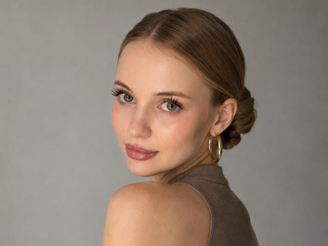 LawanaDavito with brown hair in a bun, green eyes, and pink lipstick, wearing a brown sleeveless top, gold hoop earrings, and looking over her shoulder with a subtle smile.