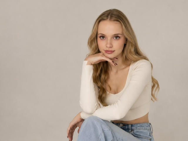 LawanaDavito with blonde hair, wearing a white long-sleeve crop top, sitting, blue jeans, hand under chin, looking at camera, soft smile, light skin, medium tits.