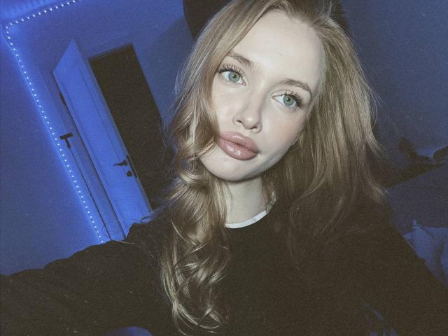 LawanaDavito with long blonde hair, green eyes, and pouty lips in a black top, taking a selfie in a blue-lit room. Her tits are not visible.