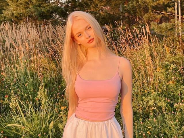 LawanaDavito with long blonde hair, pink tank top, white skirt, small boobs, slim, standing in tall grass, sunlight, green background, calm expression, natural light, outdoor setting.