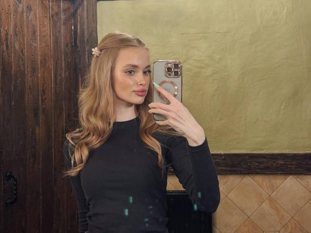 LawanaDavito with long blonde hair, pink flower hairpin, black long-sleeve shirt, holding phone in mirror, reflection showing phone case with ring. Her tits are small, she's cute.
