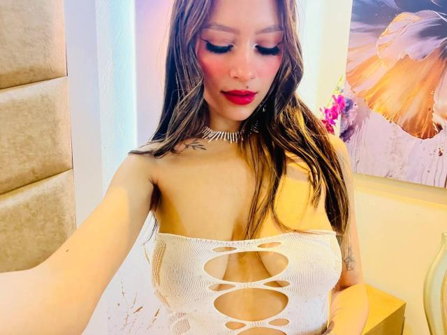 AfroditaWild with long brown hair, red lipstick, and white cut-out top showing her tits, wearing a sparkling choker, looking down with a slight smile.