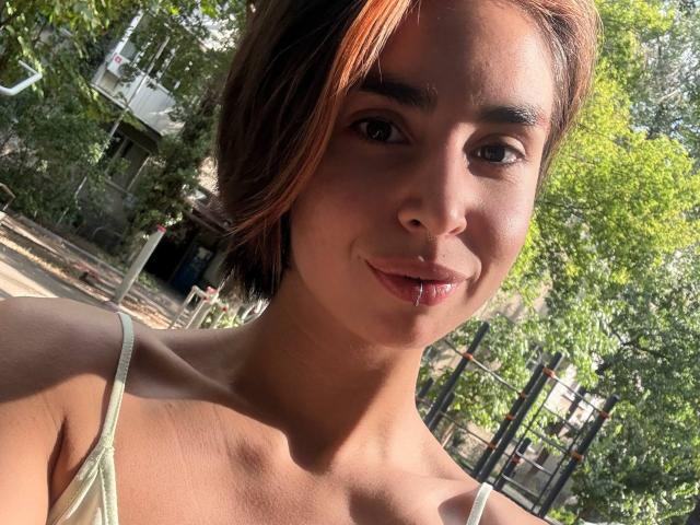 TameikaMacclellan has light brown skin, short brown hair with red highlights, small nose, and is smiling with a drop of saliva on her lower lip, wearing a green top.