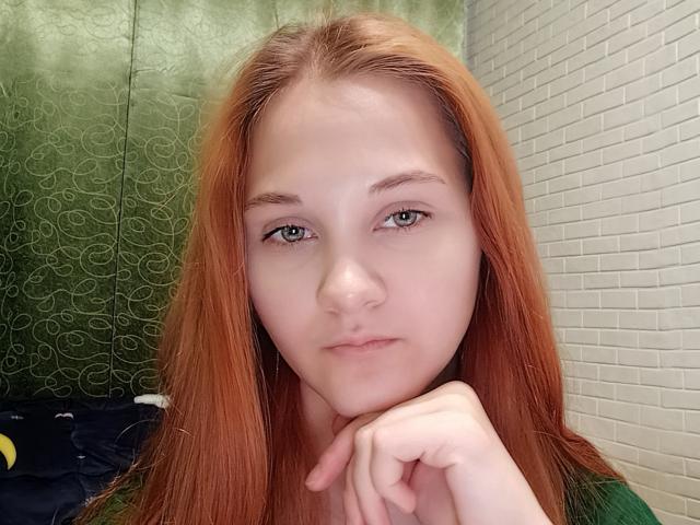 CarissaCariddi has long red hair, pale skin, green eyes, and a small nose. She's resting her chin on her hand, looking straight at the camera with a neutral expression.