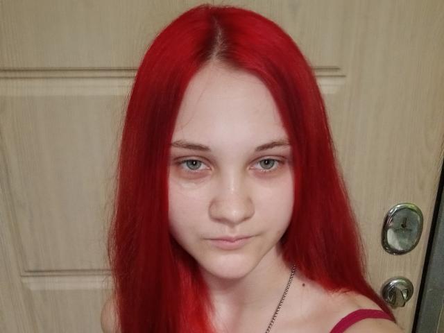 CarissaCariddi with red hair, pale skin, green eyes, and a slight smile, wearing a pink strap, standing in front of a wooden door with a silver handle.