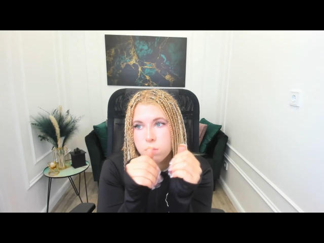 SaraMuger with blonde braids, blue eyes, and a pouty face, giving a thumbs-up with her right hand, wearing a black long-sleeve shirt.