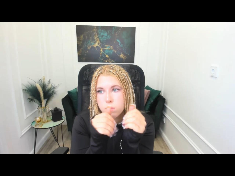 SaraMuger with blonde braids, blue eyes, and a pouty face, giving a thumbs-up with her right hand, wearing a black long-sleeve shirt.