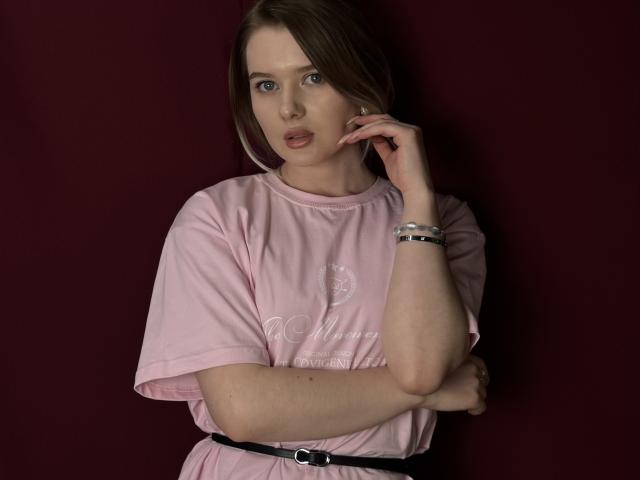 Cynthia Milkowski con una camicia rosa pallido, una cintura nera e un braccialetto d'argento, la mano vicino al viso, capelli castani, sfondo rosso scuro, con un'espressione leggermente sorpresa, con piccoli seni visibili..