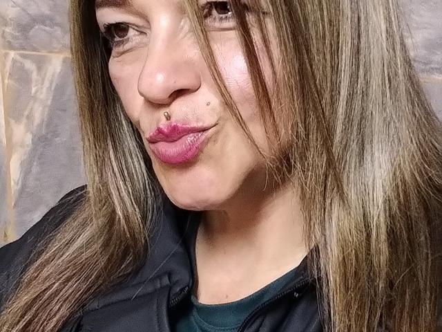 CarolMissx with long brown hair, pink pouty lips, nose ring, and a black jacket, showing off her cute face and slightly flushed skin in a selfie.