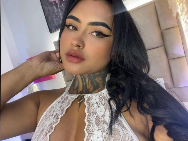 LairaHearvy with long black hair, dark eyebrows, and thick eyeliner, wearing a white lace halter top, showing a tattooed neck and pink lips, looking at the camera.