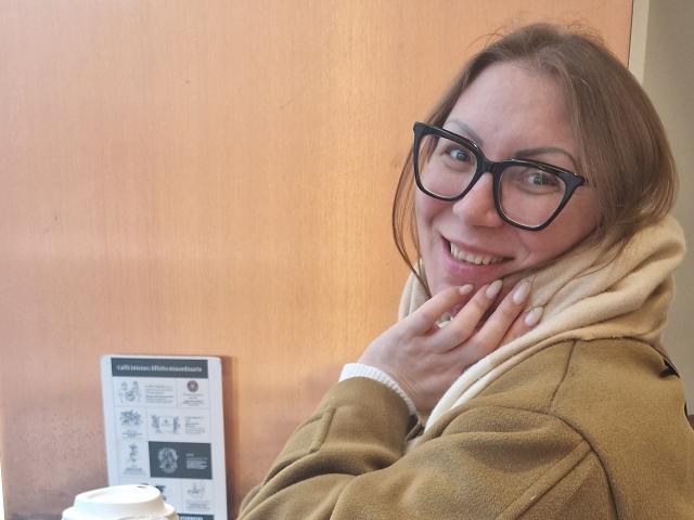 ErikaMilf with glasses, brown hair, smiling, wearing a beige coat and white scarf, hand near mouth, showing a ring, wooden door in background.