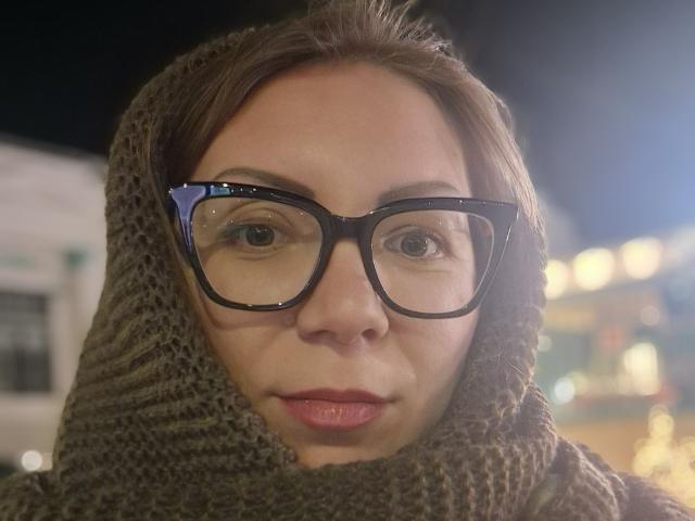 ErikaMilf with glasses, brown hair, and a knit scarf, has a serious expression and light skin. Boob-free, no ass or pussy visible. Night setting with blurry lights.