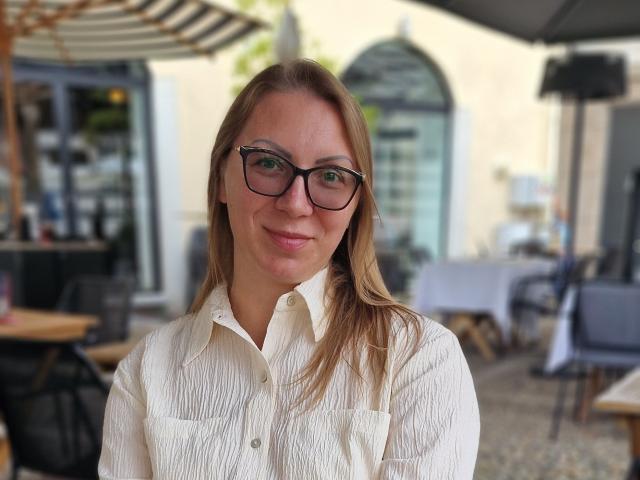 ErikaMilf with blonde hair, glasses, and a white button-up shirt, smiling slightly, showing her small tits. Blurred outdoor café in the background.