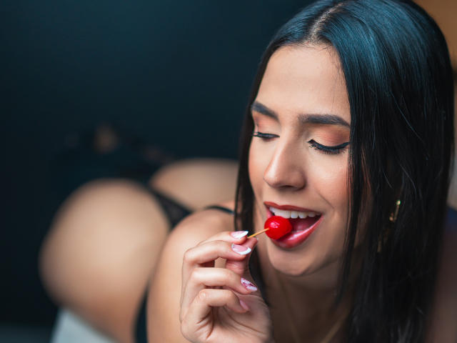 Ivory100, con i capelli neri, il rossetto rosso e gli occhi chiusi, succhia una caramella rossa, mostrando le unghie bianche, indossando biancheria intima nera ed esponendo il suo sedere rotondo in una posa sensuale..