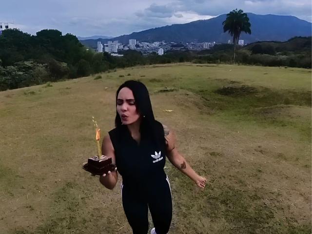 JessicaFiore, con lunghi capelli neri, indossa una canotta nera senza maniche con un logo bianco, tiene in mano una tazza marrone con una cannuccia arancione, è in piedi su una collina erbosa, con montagne e una città sullo sfondo, e un cielo nuvoloso..