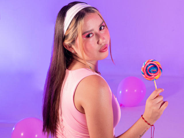 Amily Rizzo mit langen braunen Haaren, einem weißen Stirnband, einem pinken Oberteil, die einen bunten, spiralförmigen Lutscher hält, mit pinken Ballons, pinker und violetter Beleuchtung, die über die Schulter schaut und dabei ihre Brüste zeigt, mit einem süßen Gesicht..