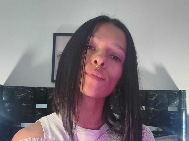 AmelieBlake has black straight hair, a nose ring, and a white sleeveless top, with a subtle smile, looking at the camera. Her face is slightly flushed.