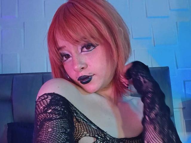 ViolettClay with red hair, black lace gloves, and dark lipstick, showing off her tits and looking seductive with a hand on her shoulder.