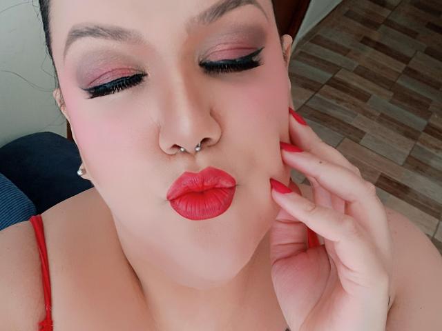 GiiannaMorrys with pink eyeshadow, black eyeliner, red lipstick, nose piercing, and red fingernails, pouting with her hand on her cheek, wearing a red strap.