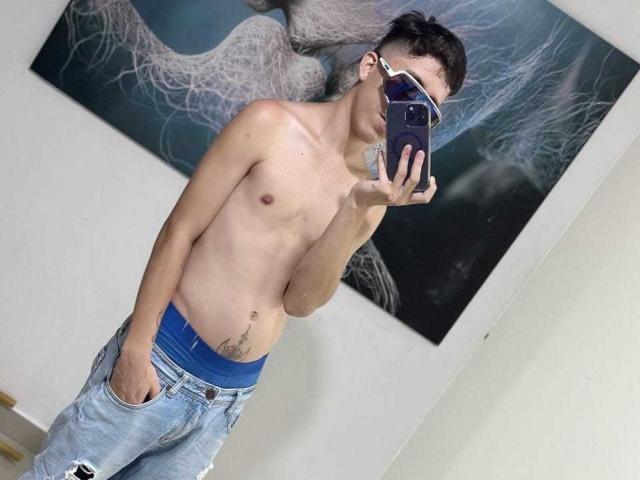 Shirtless SantiagoColeman with a slim, toned body, wearing blue jeans and a blue waistband, holding a phone in front of his face, showing a tattoo on his right side.
