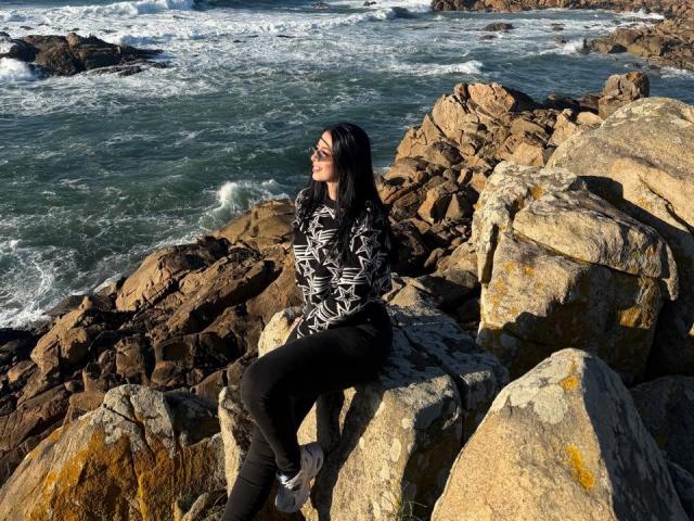 IsabellaJames with black hair and sunglasses, wearing a black and white patterned top and black pants, sitting on rocky shoreline, looking at the ocean.