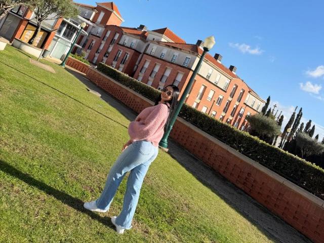 IsabellaJames stands on grass with a slanted camera angle, wearing a pink shirt, blue jeans, and white shoes, looking back with a short beard and dark hair.