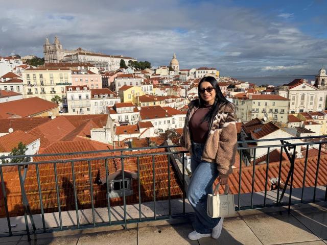 IsabellaJames with long dark hair, sunglasses, brown jacket, and blue jeans, standing on a balcony with red rooftops, holding a white bag, looking over a city.