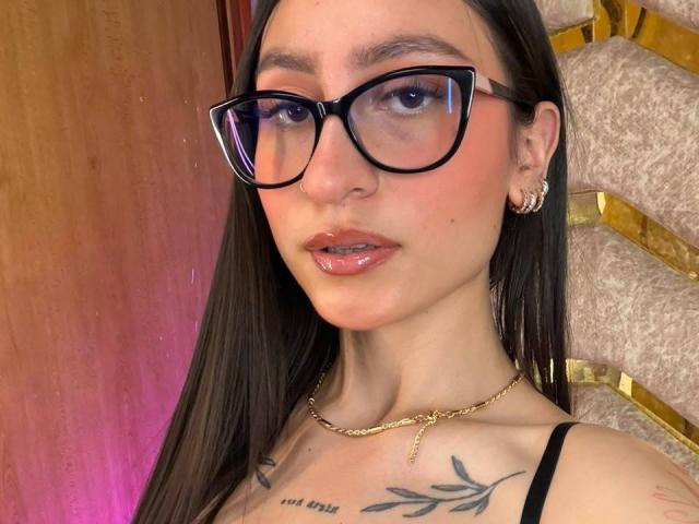 MaddyCarina heeft een bril, glanzende roze lippen en lang donker haar. Ze draagt een zwarte top met bandjes, een gouden ketting en oorbellen. Ze heeft tatoeages op haar borst. Haar huid is enigszins roze, met subtiele make-up..