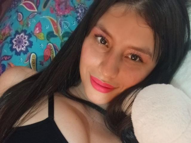 MadissonFontaine tem cabelos longos e escuros, batom rosa e um sorriso adorável. Ela está vestindo uma blusa preta, que mostra seus seios, e está deitada em um travesseiro colorido com estampa floral..