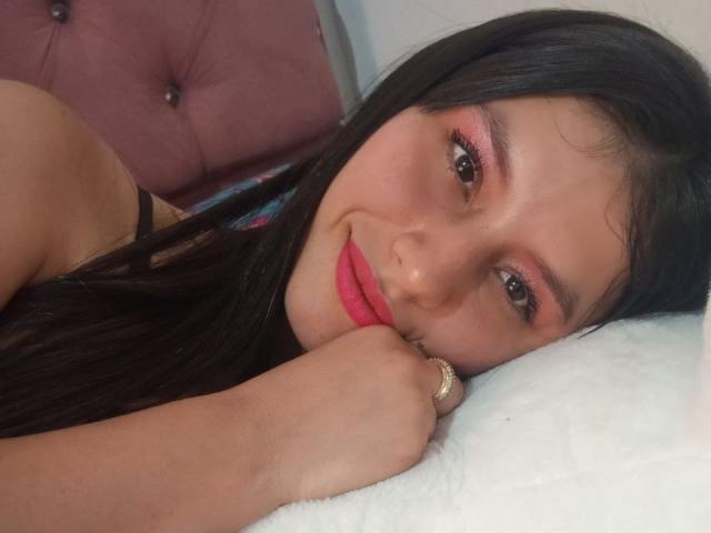 MadissonFontaine's face, laying on a white pillow, with pink eyeshadow, red lipstick, and black hair, smiling, hand under chin, wearing a ring, pink tufted headboard in background.