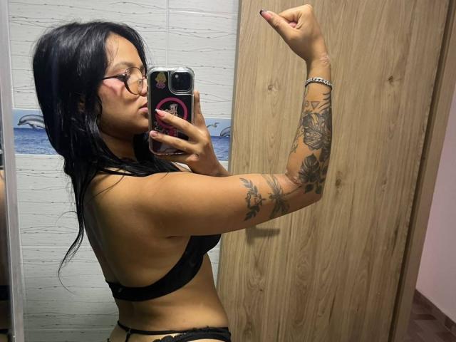 AlanaLuxe met zwart haar, een bril en tatoeages, die een spiegelselfie maakt in een zwarte bh, waarbij haar zijkant te zien is en ze naar de camera wijst, met een houten deur op de achtergrond..