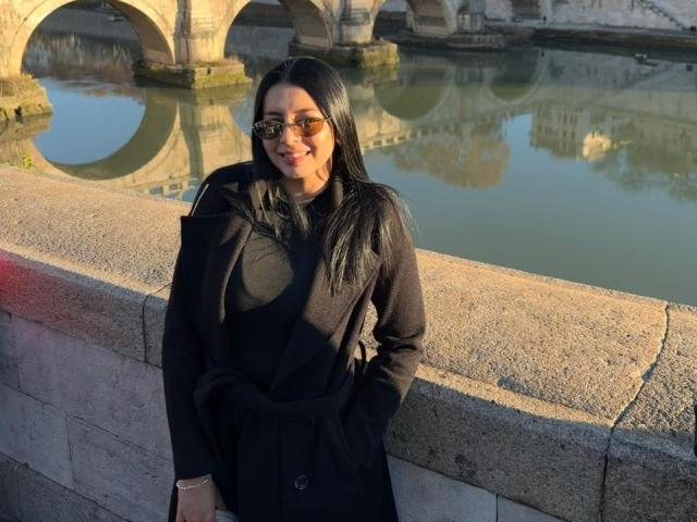 IsabellaJames with long black hair, wearing a black coat and black top, standing by a stone wall, smiling, hands in pockets, wearing glasses, reflection in water.