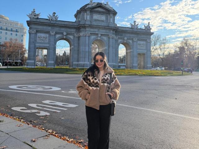 IsabellaJames stands in front of a gray stone archway, wearing a beige and black chevron-patterned jacket, black pants, and sunglasses, with hands in pockets, holding a black purse.