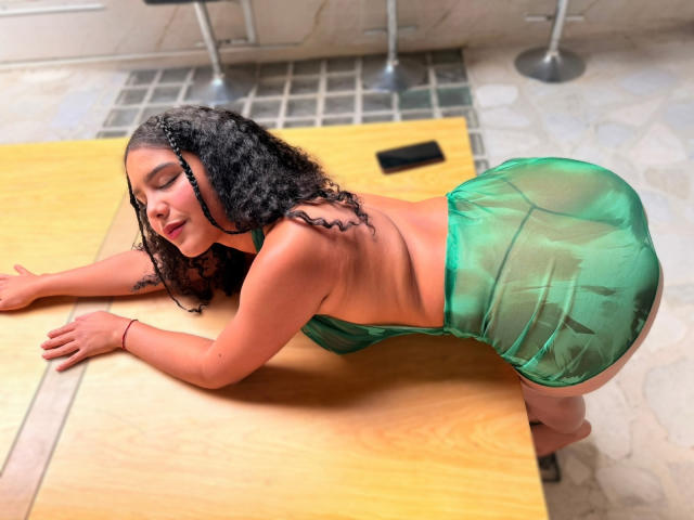 AriaVega, with curly black hair, wearing a green sheer dress, is bent over a wooden table, showing her ass, with a red bracelet on her right wrist.