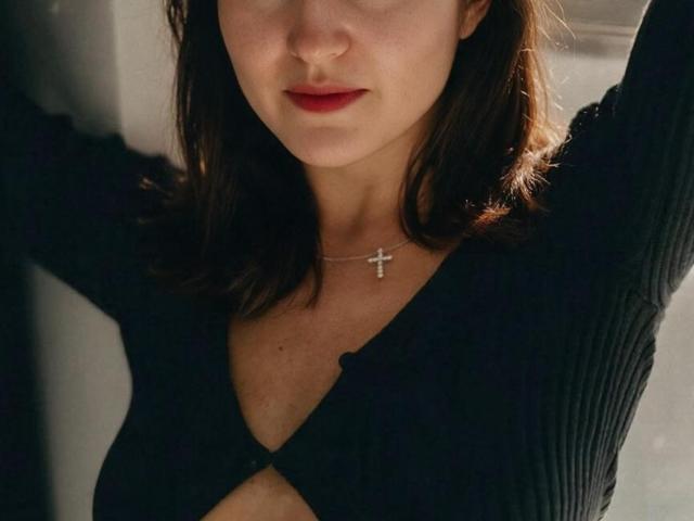 RosalindCutsinger with red lips, black ribbed top, arms raised, cross necklace, light brown hair, slight shadow, soft light, visible cleavage.