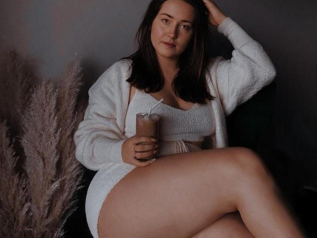 RosalindCutsinger mit mittelgroßer Brust, trägt einen weißen, flauschigen Cardigan und Shorts, hält ein Glas Eiskaffee, sitzt mit einem Bein angezogen, hat dunkelbraunes Haar und einen neutralen Gesichtsausdruck..