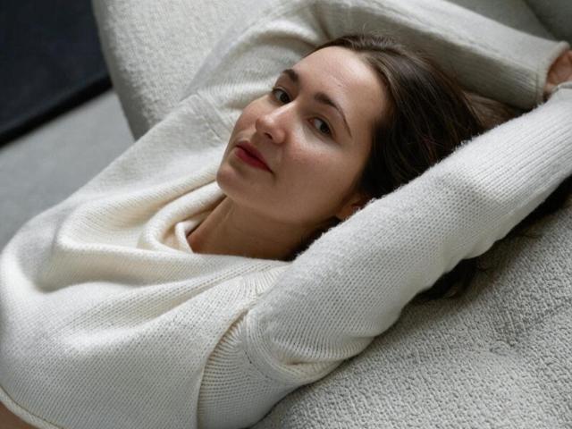 RosalindCutsinger mit erhobenen Armen, liegend auf einem strukturierten grauen Sofa, trägt einen weißen Strickpullover, hat braunes Haar, roten Lippenstift und einen entspannten Gesichtsausdruck..