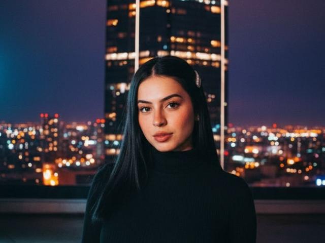 IsabellaJames with long black hair, wearing a black turtleneck, has a serious look, standing in front of a city night skyline with a tall building in the background.