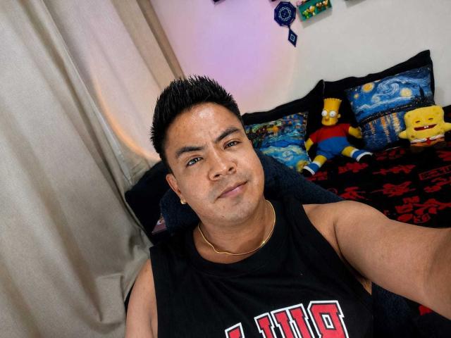 RagoForYou has a chubby face, short black spiky hair, wearing a black "HBO" tank top, gold necklace, and a slightly confused expression, sitting on a dark blue bed.