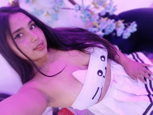 GemmaBellini with long dark hair, pink-lit skin, large tits in white strapless top, looking at camera, hand on hip, floral decorations in background.