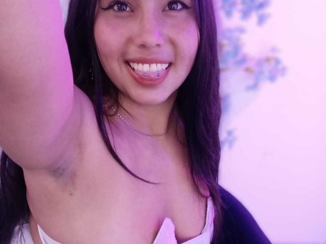 GemmaBellini with long black hair, smiling, showing tongue piercing, lifting arm, exposing armpit, white strap top, pink-purple light, visible cleavage, small nose ring, silver necklace.