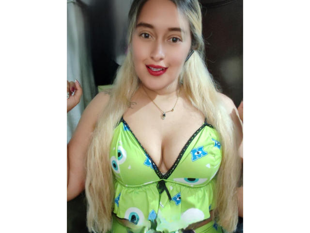 Ckamilaa69 com longas tranças loiras, blusa verde estampada, detalhes em renda preta, decote profundo, batom vermelho, sorrindo, pequena tatuagem no ombro direito, colar, seios de tamanho médio..