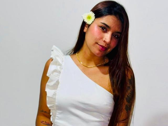 SophiaJhane with long brown hair, white dress with ruffle sleeve, gold necklace, white flower in hair, tattoos on arms, pink blush, slight smile, looking at camera.