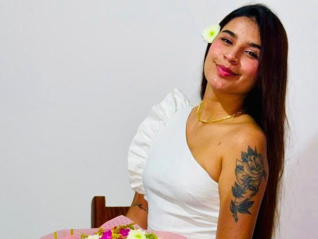 SophiaJhane with long brown hair, white flower in hair, white ruffled top, gold necklace, tattooed arm, pink skirt, smiling, sitting, pink lipstick, white wall background.