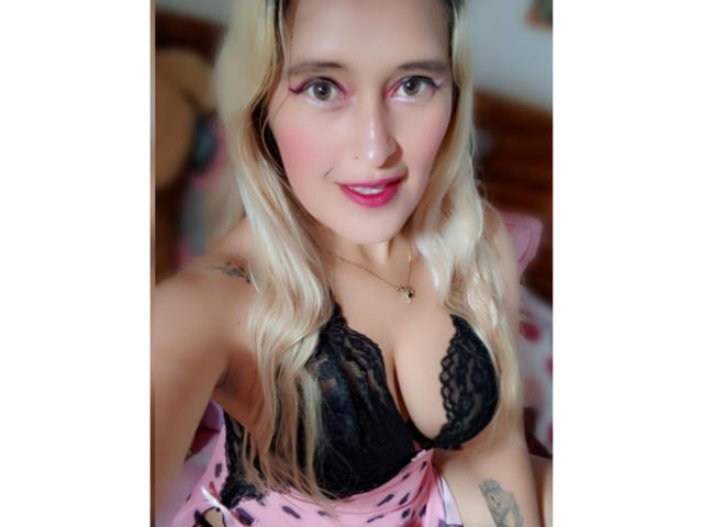 Ckamilaa69, con cabello rubio, sombra de ojos rosa y lápiz labial rosa, lleva un sujetador de encaje negro que muestra sus grandes senos, sonriendo a la cámara, y vistiendo una blusa rosa..