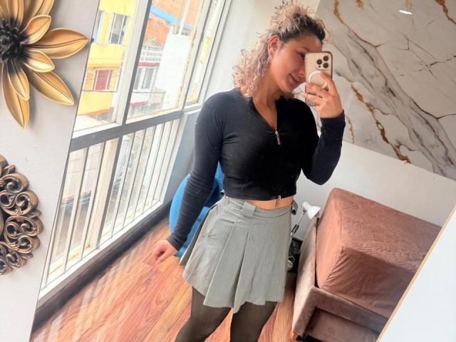 ElenaSaenz with curly pinkish hair, black long-sleeve top, gray pleated skirt, black tights, holding phone, smiling, standing in room with wooden floor, gold wall decor.
