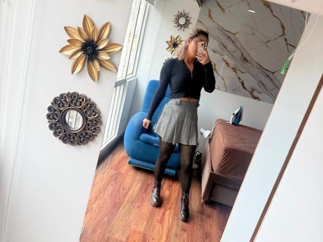 ElenaSaenz in a black crop top, grey skirt, black tights, and black boots, taking a mirror selfie, showing her curly hair and standing in a room with wooden floors.