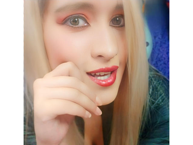 Ckamilaa69, con i capelli biondi, il rossetto rosso e l'ombretto rosa, sorride leggermente, tenendosi il mento con la mano, mostrando i denti. I suoi occhi sono verdi e sta guardando verso l'alto..