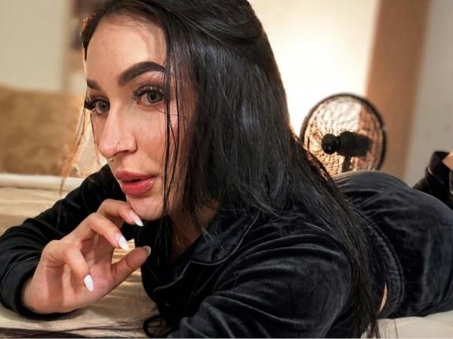 PamelaLiss, lying on her stomach, black velvet top, long black hair, white nail polish, nose ring, black pants, holding a cigarette, fan in the background, looking at camera.