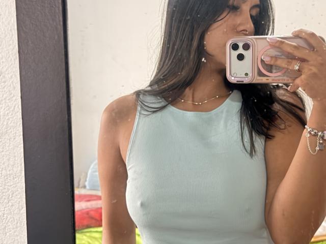 SarahPrincess with long dark hair, wearing a light blue ribbed tank top, showing her small tits, holding a pink phone, in a mirror selfie. Her wrist has a bracelet.